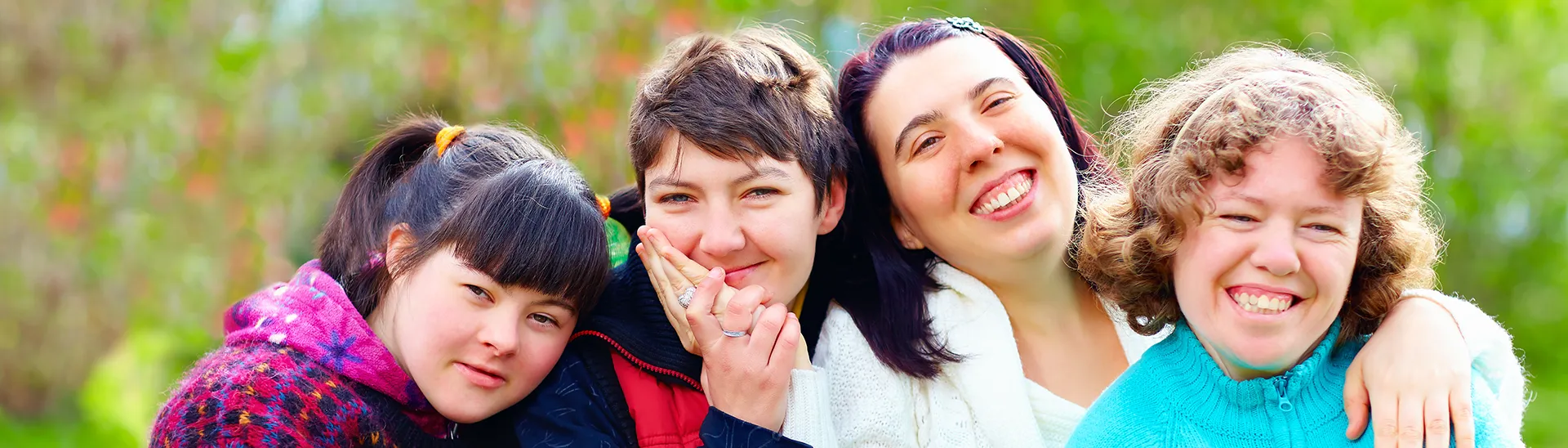 Cuatro mujeres de diferentes edades sentadas juntas, abrazadas y sonrientes en un fondo lleno de naturaleza.