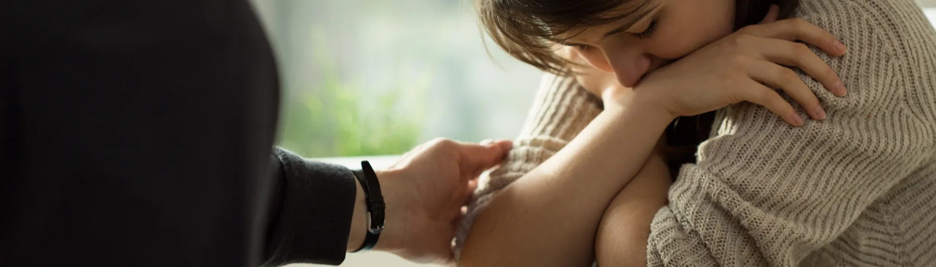 A woman in crisis with her hands folded over each other and her head down, while another person extends a comforting hand.
