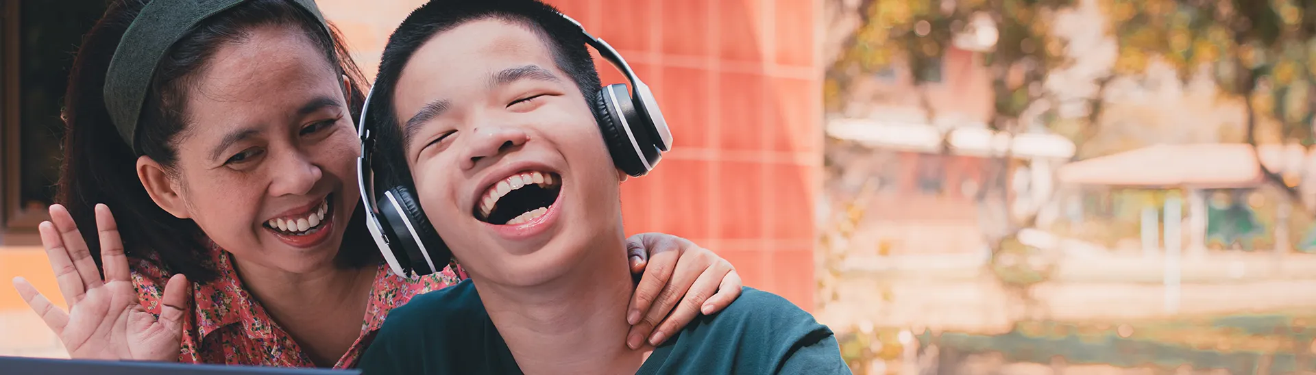 Child with down syndrome in a wheelchair wearing headphones laughing, while a woman is smiling with her hand around his neck sharing a happy moment.