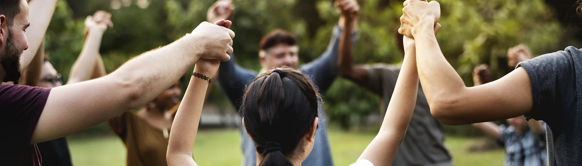Group of people with different ethnicities outside holding hands up together in a circle.