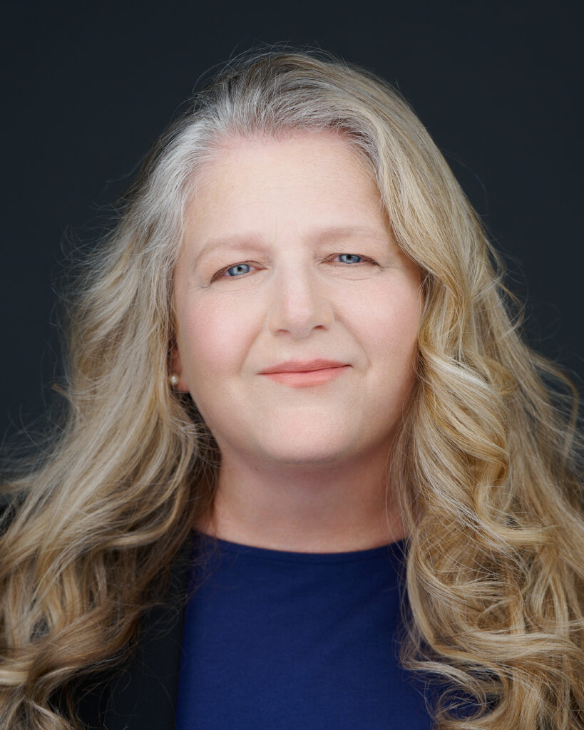 a picture of Tracy Hayes with long, wavy blonde hair is smiling slightly against a dark background, wearing a blue top and black jacket.