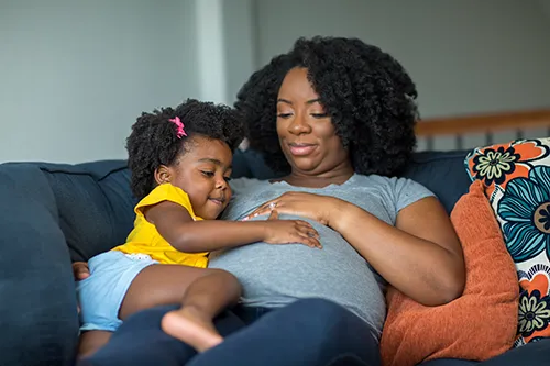A pregnant woman with curly hair is sitting on a couch, smiling warmly as her young daughter rests her head and hands on her belly. The little girl, wearing a yellow shirt, looks curious and affectionate. The cozy setting includes soft cushions, and the scene radiates love, connection, and anticipation.