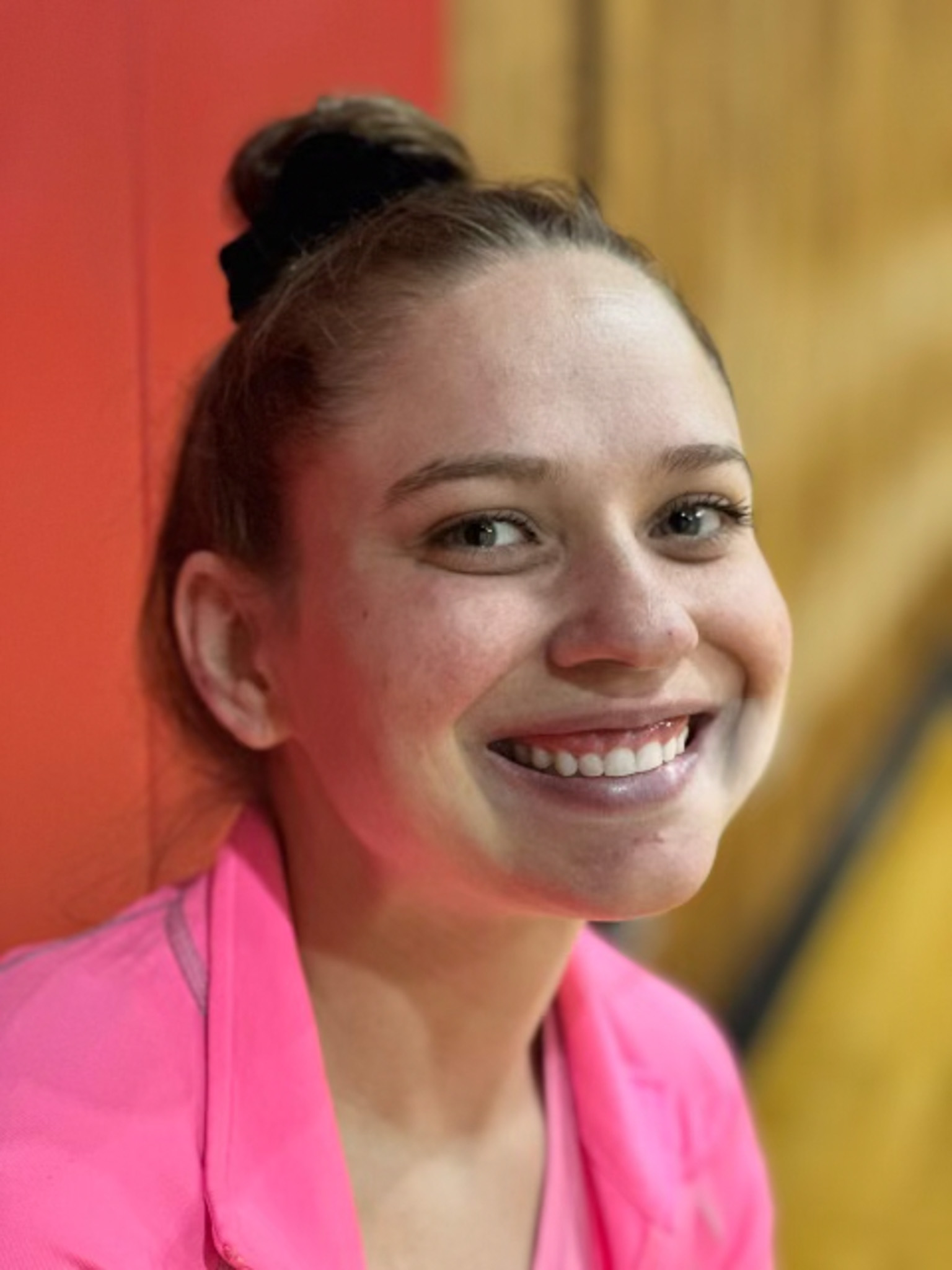 Kasi smiles widely, wearing a bright pink shirt, with a red wall in the background.