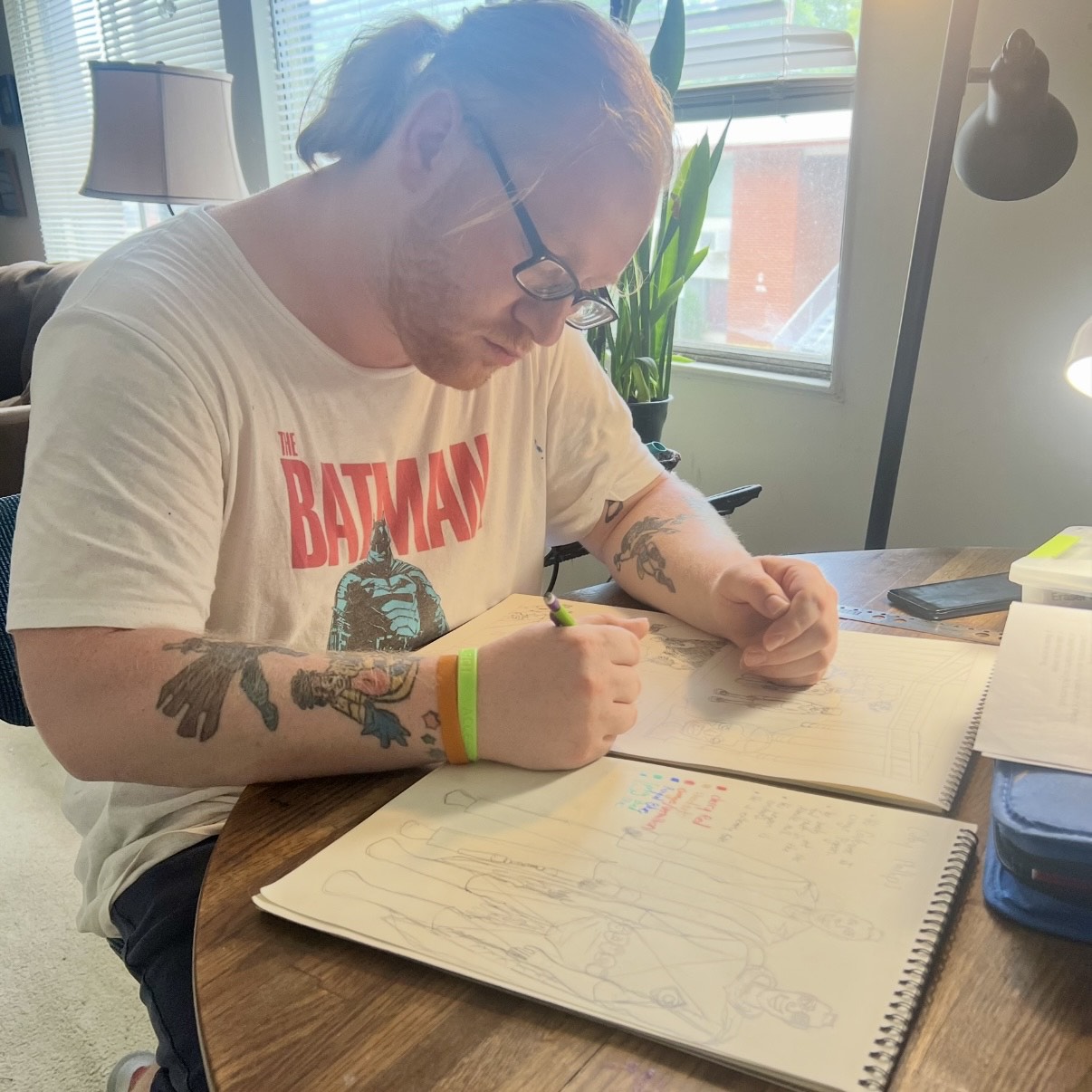 A person with tattoos, wearing a "The Batman" shirt, draws in a sketchbook at a wooden table, near a window with plants and a lamp.