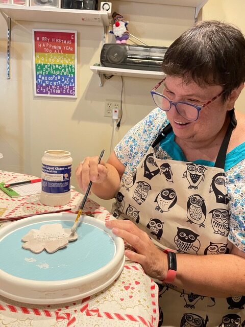A person wearing glasses and an owl apron paints a ceramic piece with glaze at a table. A colorful poster in the background reads “Merry Christmas Happy New Year.”