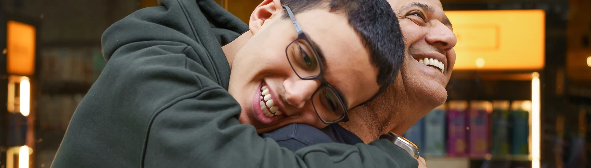 A son with glasses hugging is dad from behind while they both smile together.