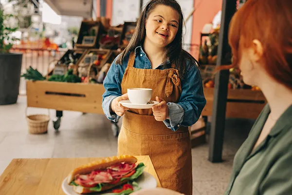Handicap-Woman-Working_2203155419 A young woman with down syndrome working and serving coffee to a woman who has a sandwich on a table in front of her.