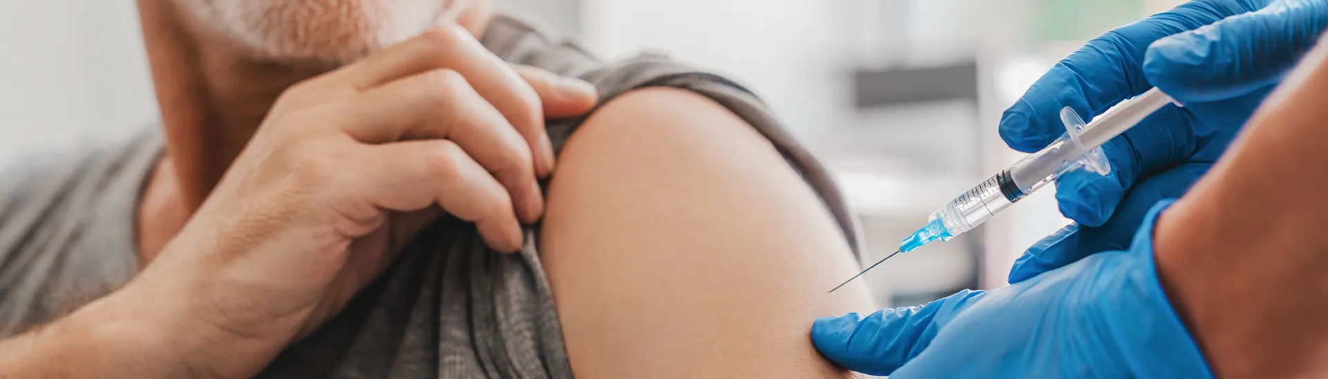 A man receives a vaccination shot in his shoulder as he holds up his sleeve while the doctor wearing blue gloves inserts the needle in his arm.