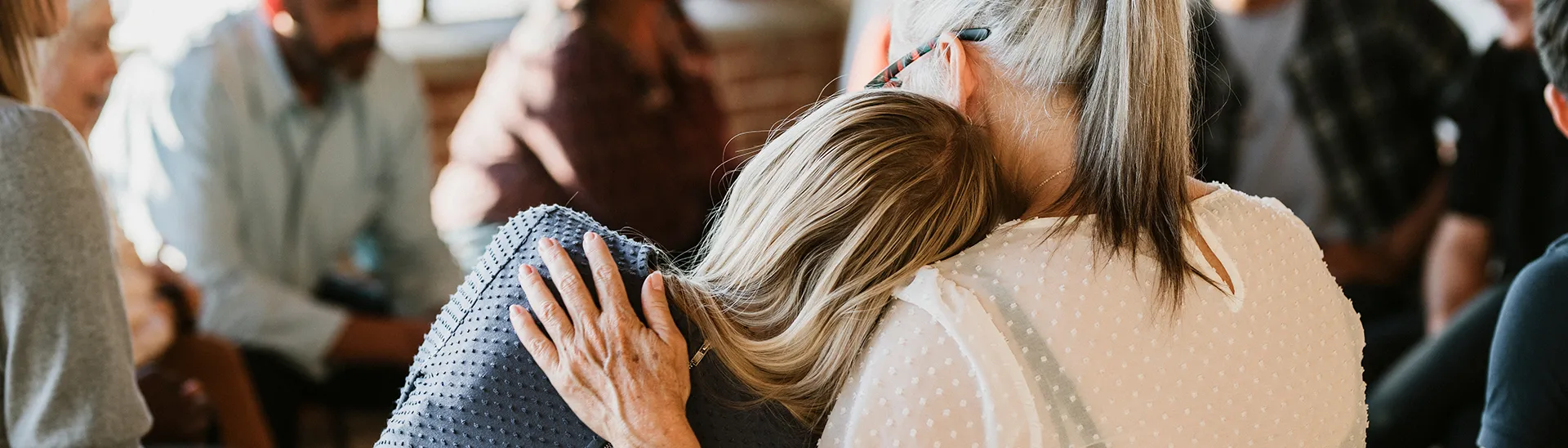 A woman rests her head on another woman for support while attending a support group with other people sitting around in a circle.