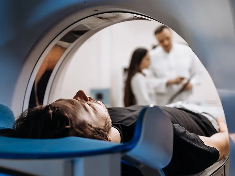 A man lays inside a CT Scan machine while two doctors talk together and review his chart.