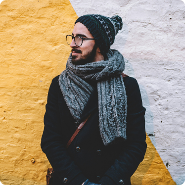 Man with glasses and scarf leaning against a concrete wall. 