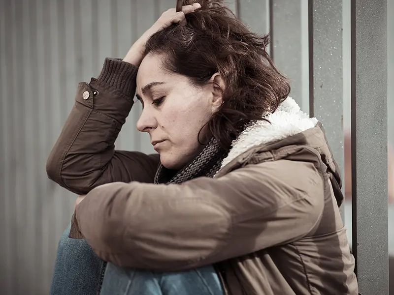 A woman with a winter coat on sitting against a rail with her hand in her hair looking distressed.